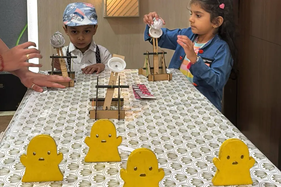 Children at a long table lined with wooden target stands and yellow ghost-shaped targets, taking aim