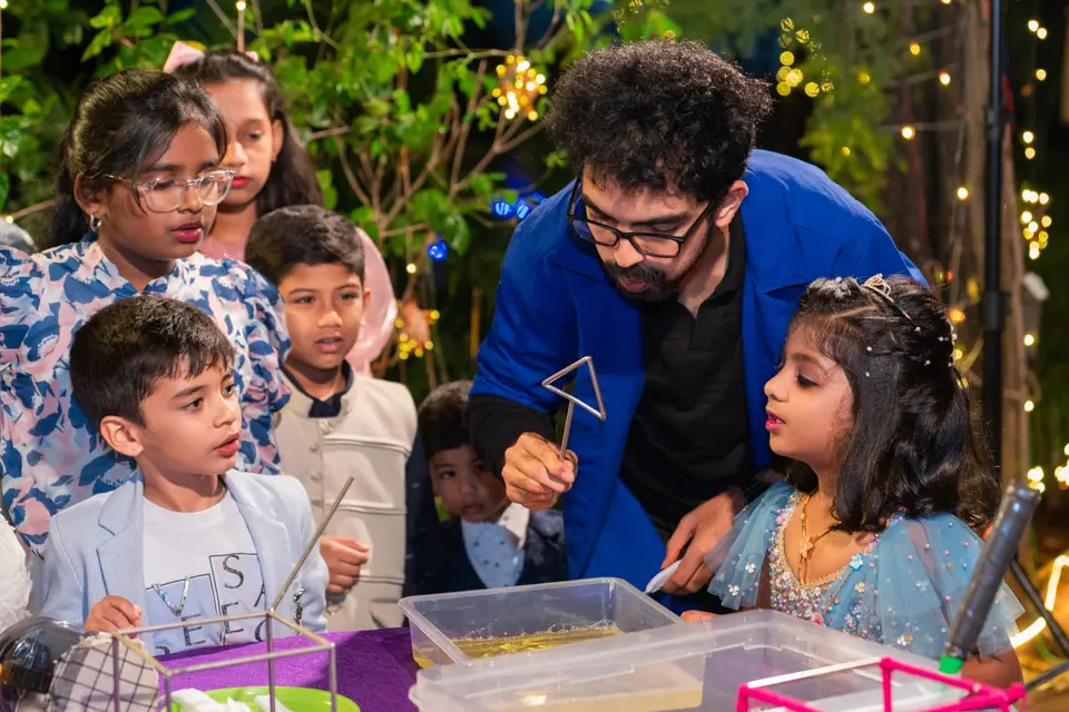 A MakerMasti host leaning over a tray of bubble solution while a crowd of children watch closely