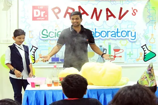A host and a child at a decorated science laboratory table as a tall column of yellow foam erupts