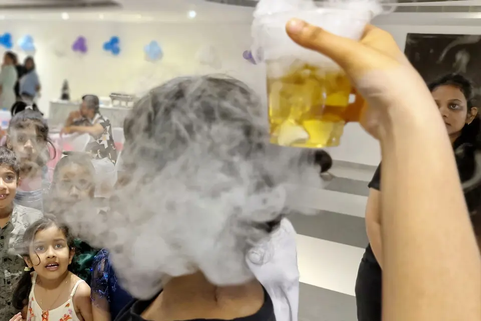 A hand tipping a jug of dry-ice fog as a row of children watch the smoke pour out over the table