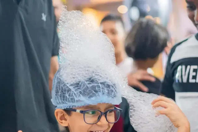 A boy laughing with a tall column of foam balanced on his head at a science birthday party
