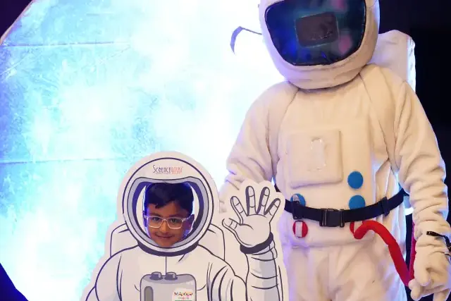 An astronaut mascot and a cut-out photo board standing in front of a large illuminated moon backdrop