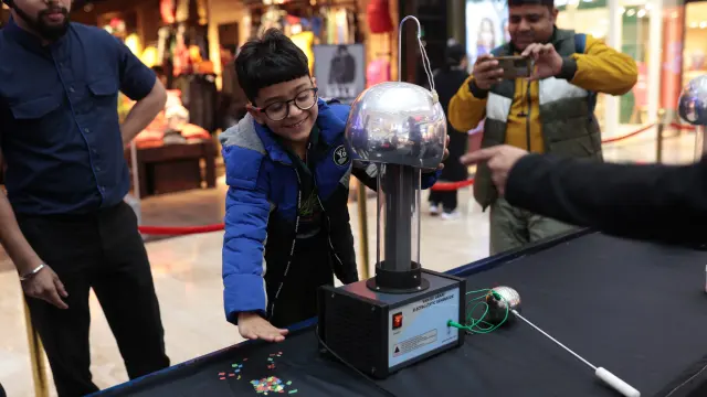 A boy grinning as he touches a Van de Graaff generator while shoppers stop to watch at a mall science festival