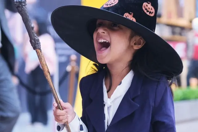 A girl in a witch hat laughing as she raises a light-up wand